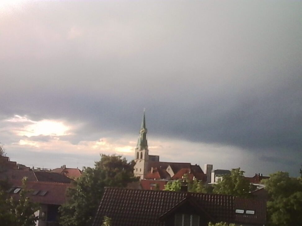 Church tower above rooftops under dark clouds with sunlight breaking through