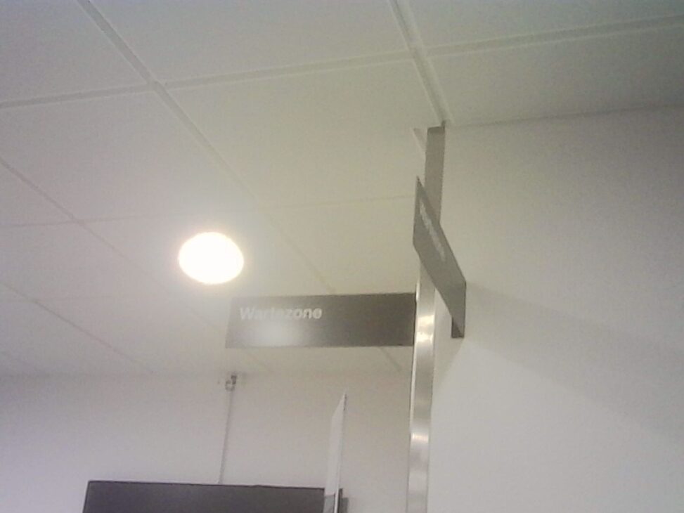 Hospital corridor ceiling with a glowing light and a hanging sign reading “Wartezone.”