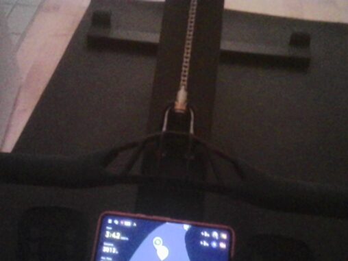 View from the seat of a rowing machine, with a smartphone mounted above the flywheel and the chain extending into the dimly lit room.