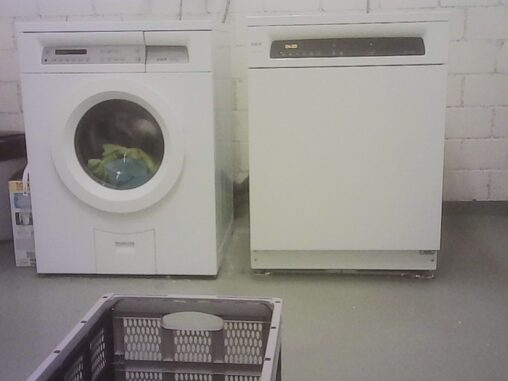 Shared laundry room in an apartment building basement, with a washing machine, dryer, and laundry basket in the foreground.