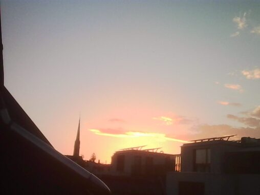 Summer sunset over rooftops, with a church spire silhouetted against a glowing orange horizon and a clear evening sky.