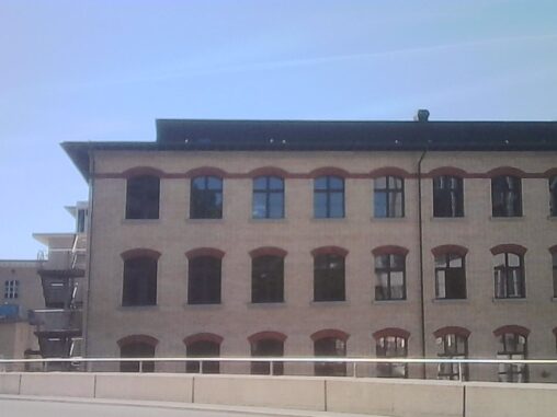 Sunlit historic brick building with rows of arched windows beneath a cloudless blue summer sky.