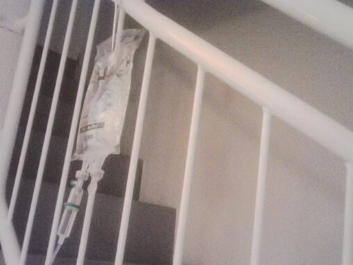 Intravenous infusion bag hanging from a white staircase railing inside an apartment during a home medical visit.
