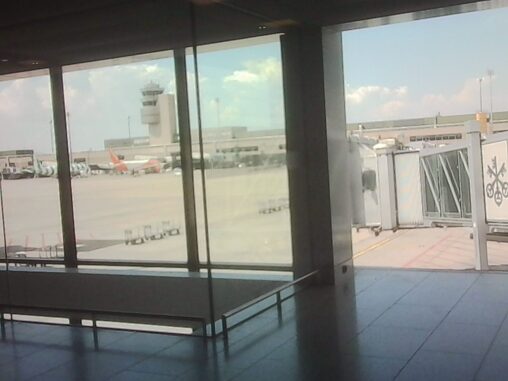 View from an airport terminal overlooking the apron, with aircraft, a jet bridge, and the control tower visible through large windows.