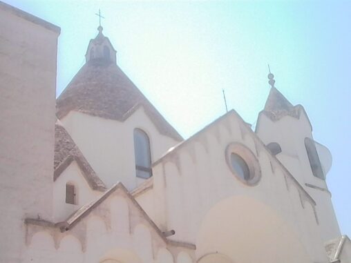 Upward view of a white church with conical roofs and bell towers beneath a bright blue summer sky in Alberobello.