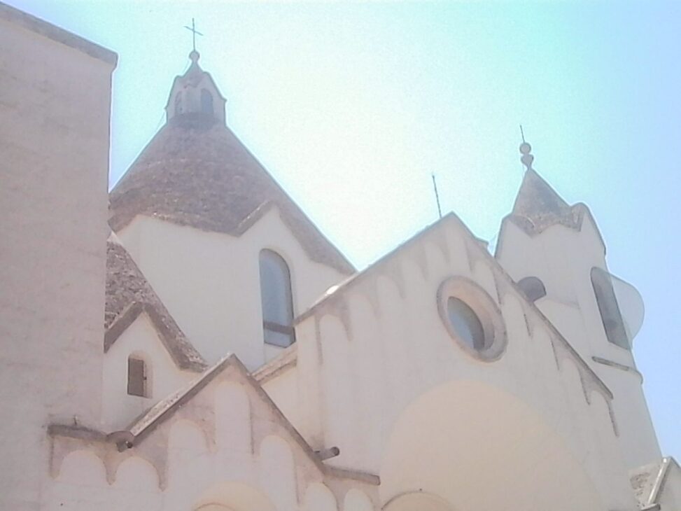 Upward view of a white church with conical roofs and bell towers beneath a bright blue summer sky in Alberobello.