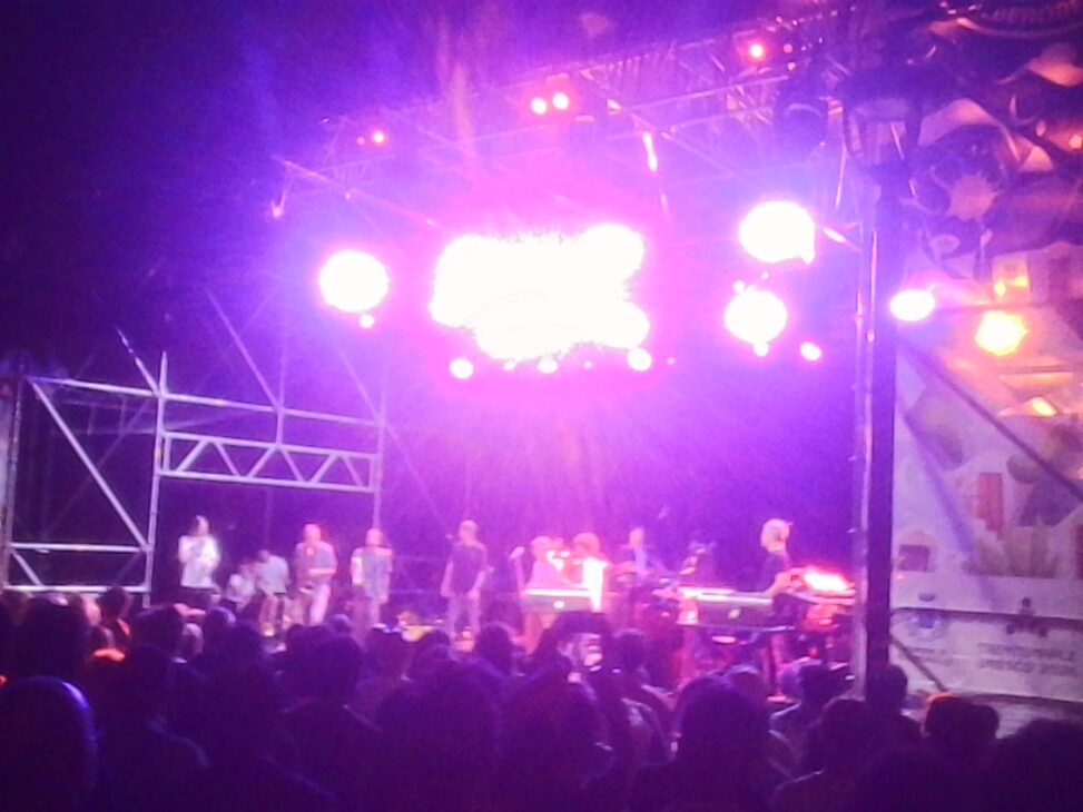 A nighttime outdoor concert with Snarky Puppy performing under vivid purple stage lights as a large audience watches from in front of the stage.
