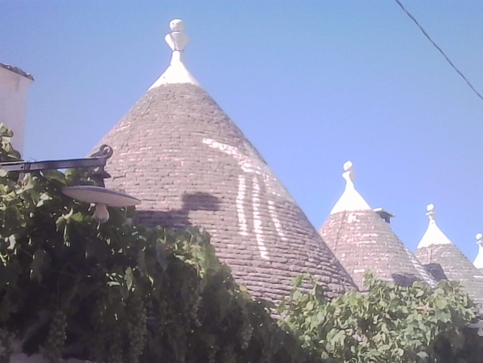 A large traditional trullo dominates the foreground, with several more cone-shaped stone rooftops receding into the distance beneath a deep blue summer sky, framed by grapevines and an old street lamp.