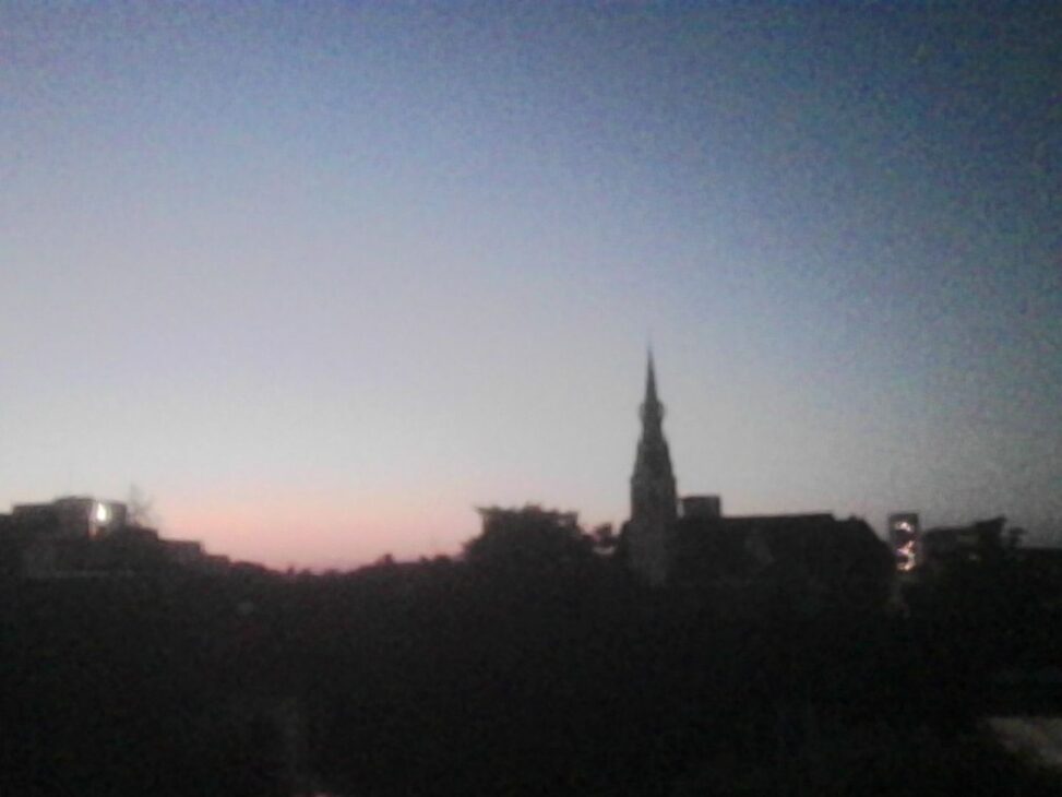 Silhouette of a church spire and the Zürich Nord skyline at dusk, with a pale pink glow on the horizon, scattered lights in distant office buildings, and a deepening blue evening sky.