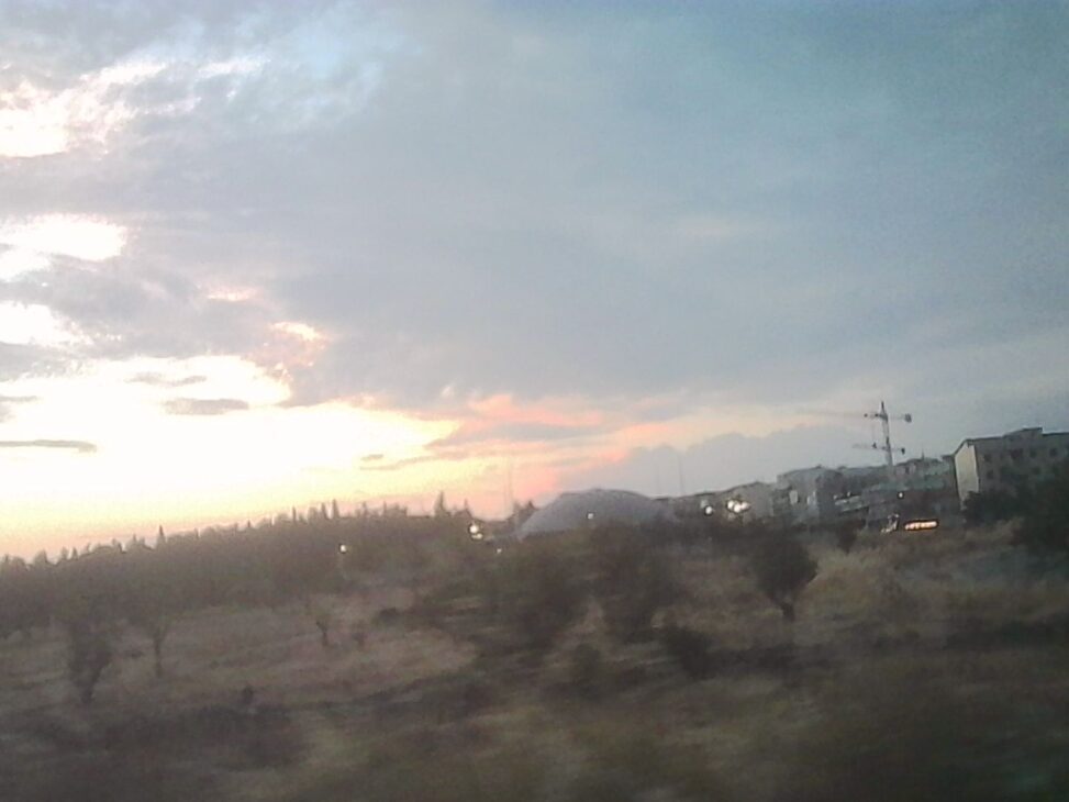 Evening view across an open field toward a weathered sports arena and nearby buildings beneath dramatic clouds glowing with the last light of sunset in Altamura.