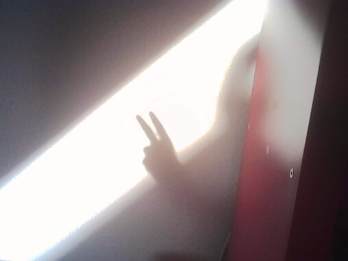 A hand making a peace sign is silhouetted against a bright beam of sunlight streaming through a partially opened window into a warm apartment.