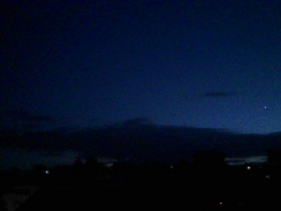 Alt text: Deep blue evening sky above dark silhouettes of trees and rooftops, with the last faint glow of sunset lingering along the western horizon and a single bright point of light visible through the twilight.