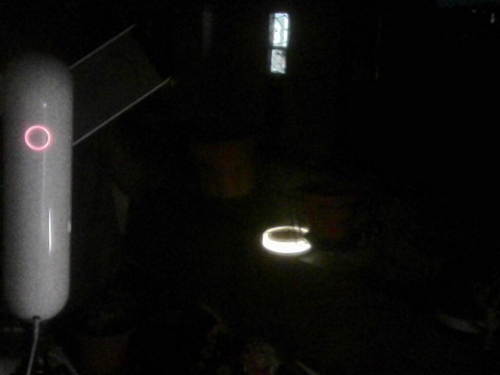 A grainy, low-light photo at night on a dark balcony. On the left, a sleek, white capsule-shaped smart telescope stands upright, its circular power button glowing with a red ring. The pitch-black background features a faint glowing white ring on the floor and a dim window in the distance, making the device resemble a sci-fi turret from the game Portal.