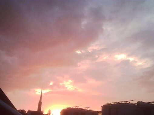 Pink and orange evening light glows beneath thick storm clouds over Zurich. A church spire and nearby rooftops form dark silhouettes against the bright horizon as the departing thunderstorm leaves behind a calmer sky.
