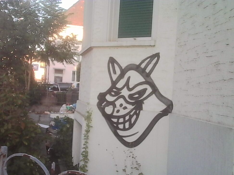 Black-and-white graffiti of a grinning, fox-like masked face painted on the corner of a white building during golden hour. Warm evening light illuminates the wall, while bicycles, overgrown plants, and neighboring houses in the background hint at an older residential area awaiting redevelopment.