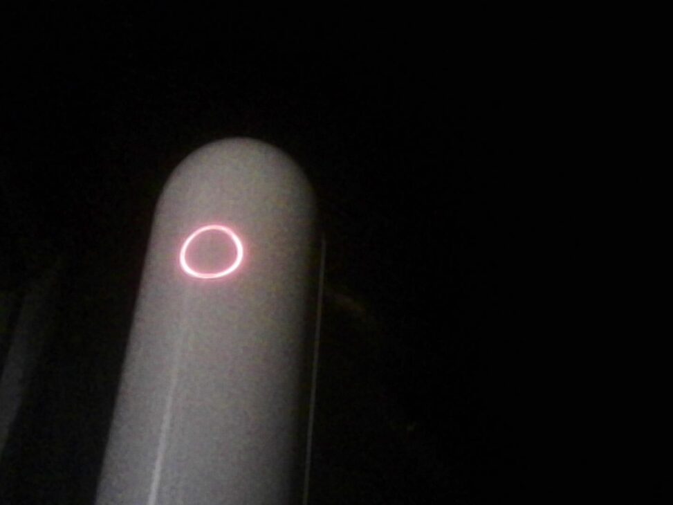 A Vespera smart telescope standing in near-total darkness, its cylindrical body faintly visible and a pinkish-red illuminated ring glowing on the front.